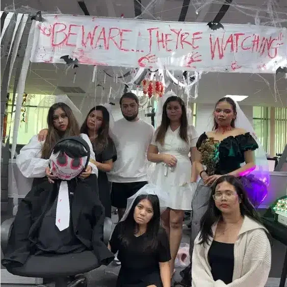 People in Halloween costumes posing in an office decorated with spooky spiderwebs and a "Beware... They're Watching" banner.
